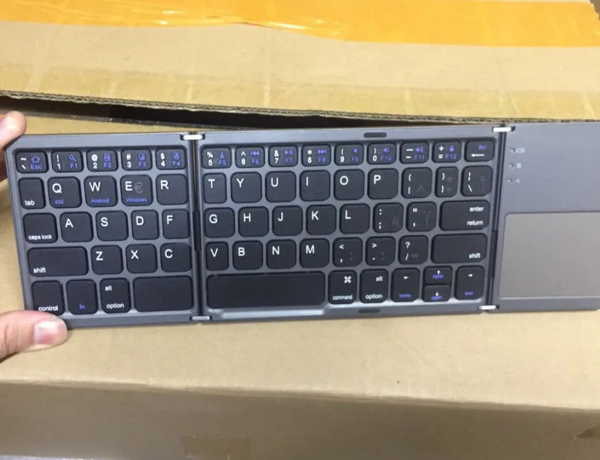 Black tri-fold portable Bluetooth keyboard with touchpad, shown folded and unfolded next to a smartphone.