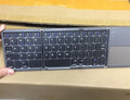 Black tri-fold portable Bluetooth keyboard with touchpad, shown folded and unfolded next to a smartphone.