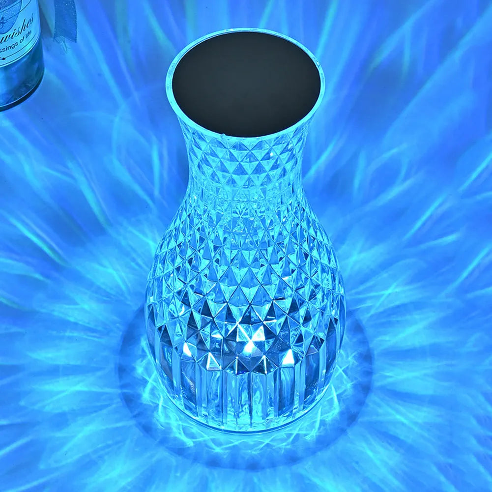 Transparent crystal vase shaped LED table lamp with remote control on a wooden bedside table.