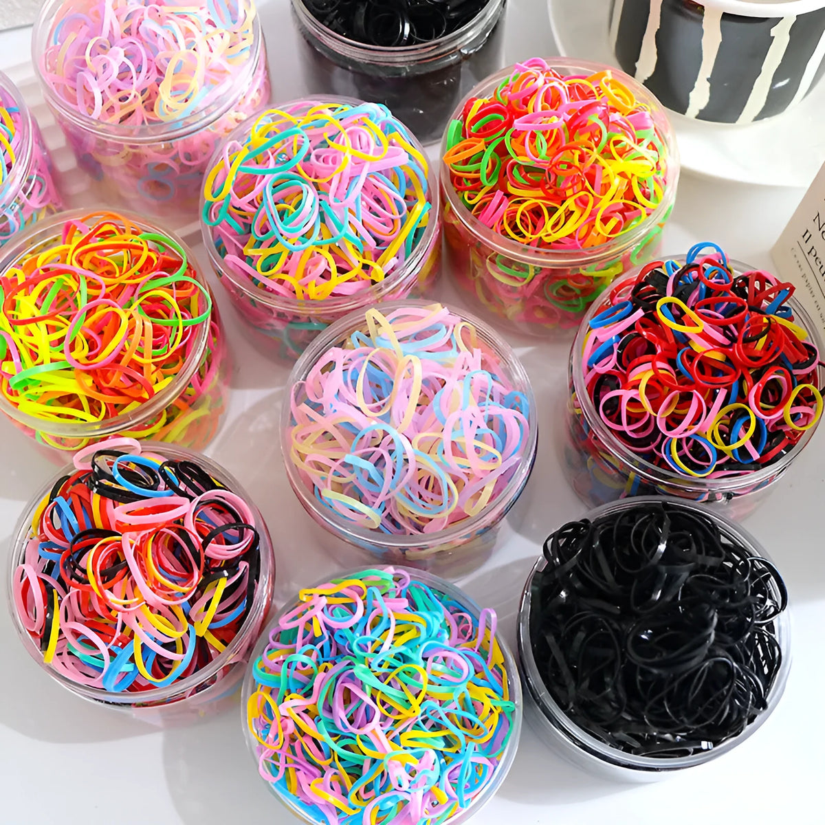 Multi-colour assortment of small TPU elastic baby hair ties in a clear reusable storage container.