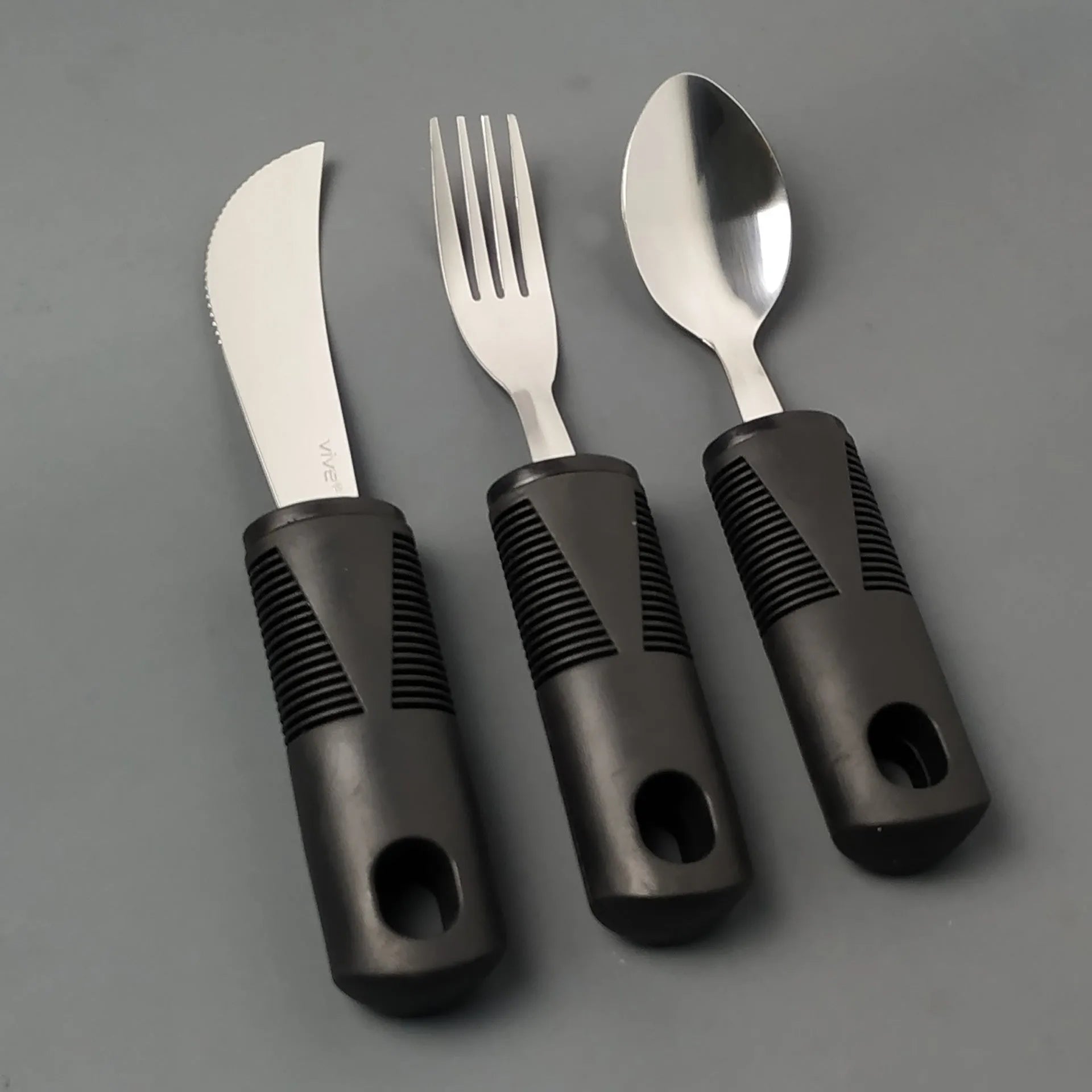 Stainless steel adaptive knife, fork, and spoon set with black rubberised easy-grip handles.