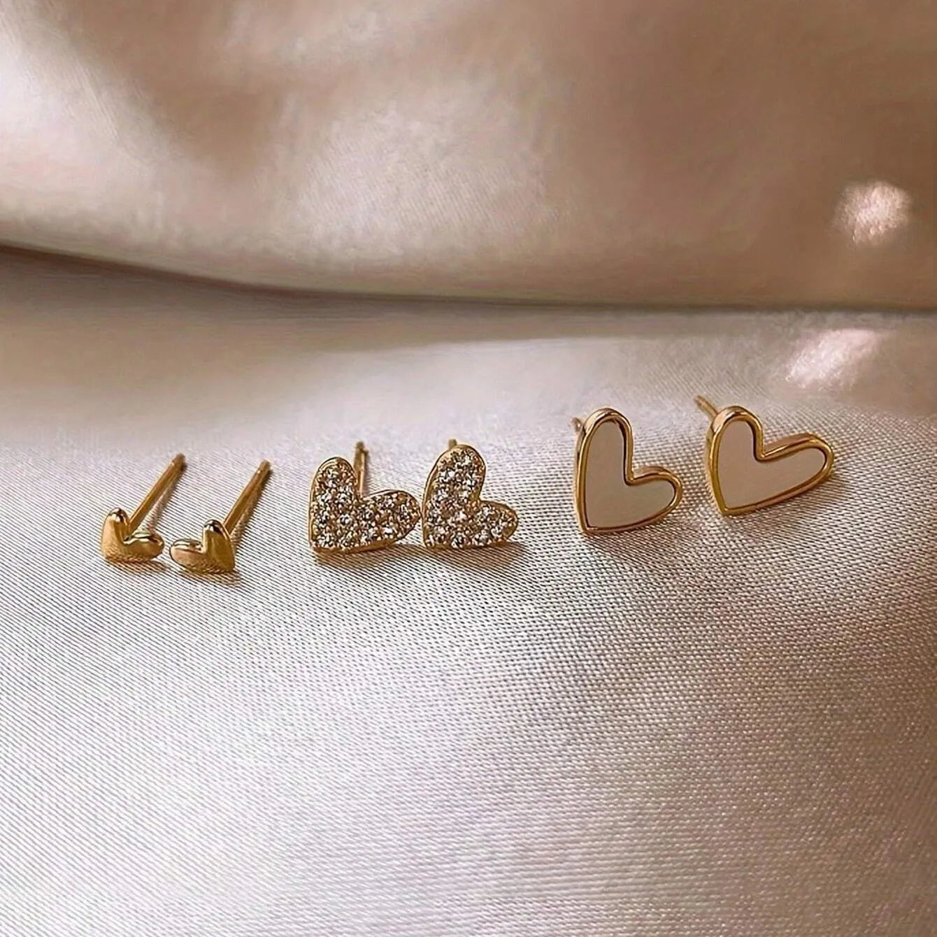 Three pairs of heart-shaped alloy earrings with electroplated finishes in gold, black, white, or pink colours.